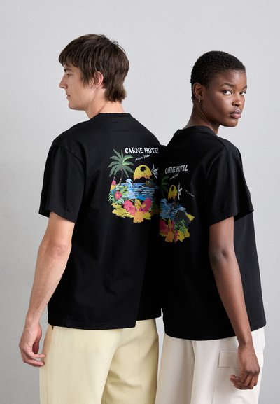 T-shirt di cotone neri indossati da due modelli, con un design grafico colorato sulla schiena che raffigura una scena tropicale e il testo "CARNE HOTEL".