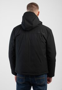 Schwarze isolierte Jacke mit Kapuze, mit glatter Oberfläche, sichtbaren Nähten und einem Logo auf dem oberen R�ücken. Wird über einer blauen Jeans getragen.