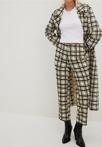 Rutig kappa och byxor i gult och svart, textil med struktur, lös passform, med en vit kort top och svarta högklackade stövlar.
