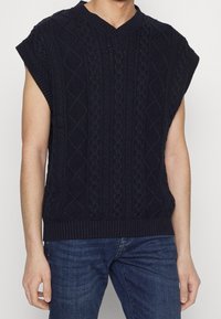 Homme portant un gilet en tricot à mailles bleu marine sans manches sur un jean bleu, se tenant avec les bras détendus le long des côtés.