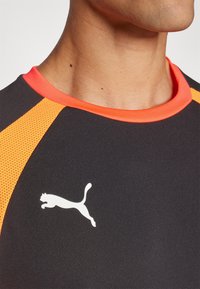 Črn atletski majica z oranžnimi mrežastimi detajli, okroglo izrezano ovratnik in belim logotipom skakajoče mačke spredaj. Gladka tekstura.