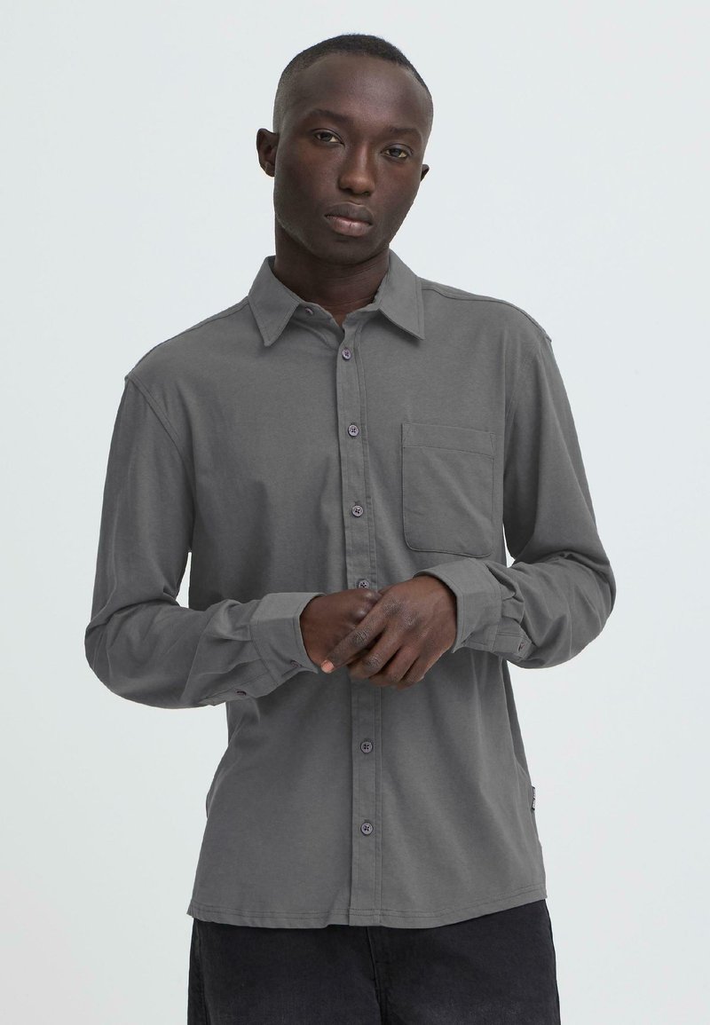 Chemise à manches longues grise en tissu léger avec un devant boutonné, une poche poitrine et des accents discrets de boutons. Design uni, sans motifs.