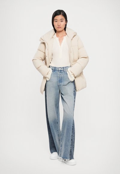 Giacca a vento di colore beige chiaro con colletto alto, polsini a coste sopra una maglietta a maniche lunghe color crema; jeans a gamba larga, blu sbiadito; sneakers bianche.