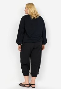 Fekete, oversize hosszú ujjú felső, elasztikus mandzsettákkal, fekete, tapered fazonú nadrággal, elasztikus szegélyekkel párosítva. Fekete lapostalpú szandálokkal viselve.