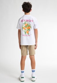 Camiseta de algodón blanca con un gráfico colorido en naranja y el texto "KEY-WEST" en la parte posterior; combinada con pantalones cortos de un beige claro y calcetines a rayas.