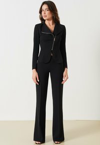 Veste noire ajustée avec fermeture éclair, forme cintrée et manches longues ; associée à un pantalon noir évasé. Tissu lisse et lignes épurées.