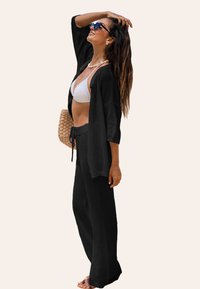 Copertura nera in maglia con maniche larghe, abbinata a un bikini bianco e pantaloni neri coordinati. Presenta una vita con coulisse e una vestibilità rilassata.