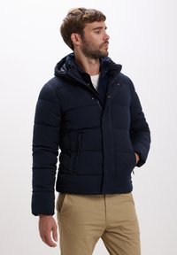 Giacca puffer blu navy con cappuccio, dotata di chiusura con zip, due tasche laterali e dettagli cuciti. Tessuto dal tocco liscio.