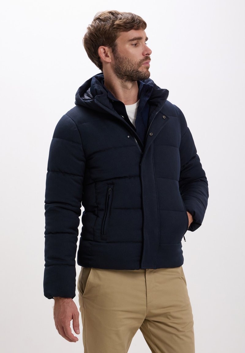 Giacca puffer blu navy con cappuccio, dotata di chiusura con zip, due tasche laterali e dettagli cuciti. Tessuto dal tocco liscio.