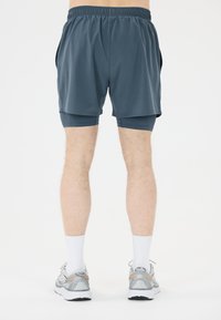 Heren sportshorts in donkerblauwe stof met een elastische tailleband. Gelaagd ontwerp met een aansluitende onderlaag, gecombineerd met lichtgrijze hardloopschoenen.
