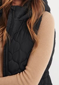 Gilet trapuntato nero con chiusura a zip, dotato di cappuccio e design testurizzato. Maglietta a maniche lunghe beige a coste che spunta da sotto.
