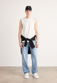 Camisa blanca sin mangas, sudadera negra atada a la cintura, jeans azul claro y zapatillas blancas. Gorra negra con logotipo. Fondo neutro.