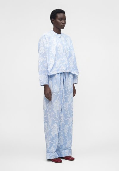 Γυναίκα που φοράει ανοιχτό μπλε μπλούζα με μακρύ μανίκι και φαρδιά παντελόνια με floral μοτίβο, συνδυασμένα με κόκκινα μαλακά παπούτσια, στέκεται μπροστά σε λευκό φόντο.