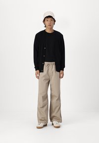 Svart knappet cardigan over en svart t-skjorte, kombinert med løse beige bukser og hvite sneakers, toppet med en lys farget strikket lue.