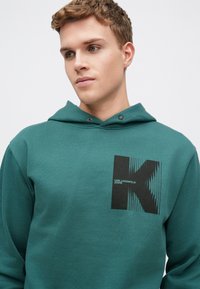 Hanorac teal din material moale, cu un logo grafic negru pe piept, design simplu, cu glugă ajustabilă și detalii minime.