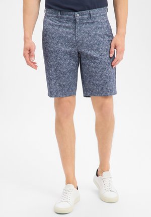 Mann trägt blaue, gemusterte kniehohe Shorts und weiße Sneaker, steht vor schlichtem Hintergrund.