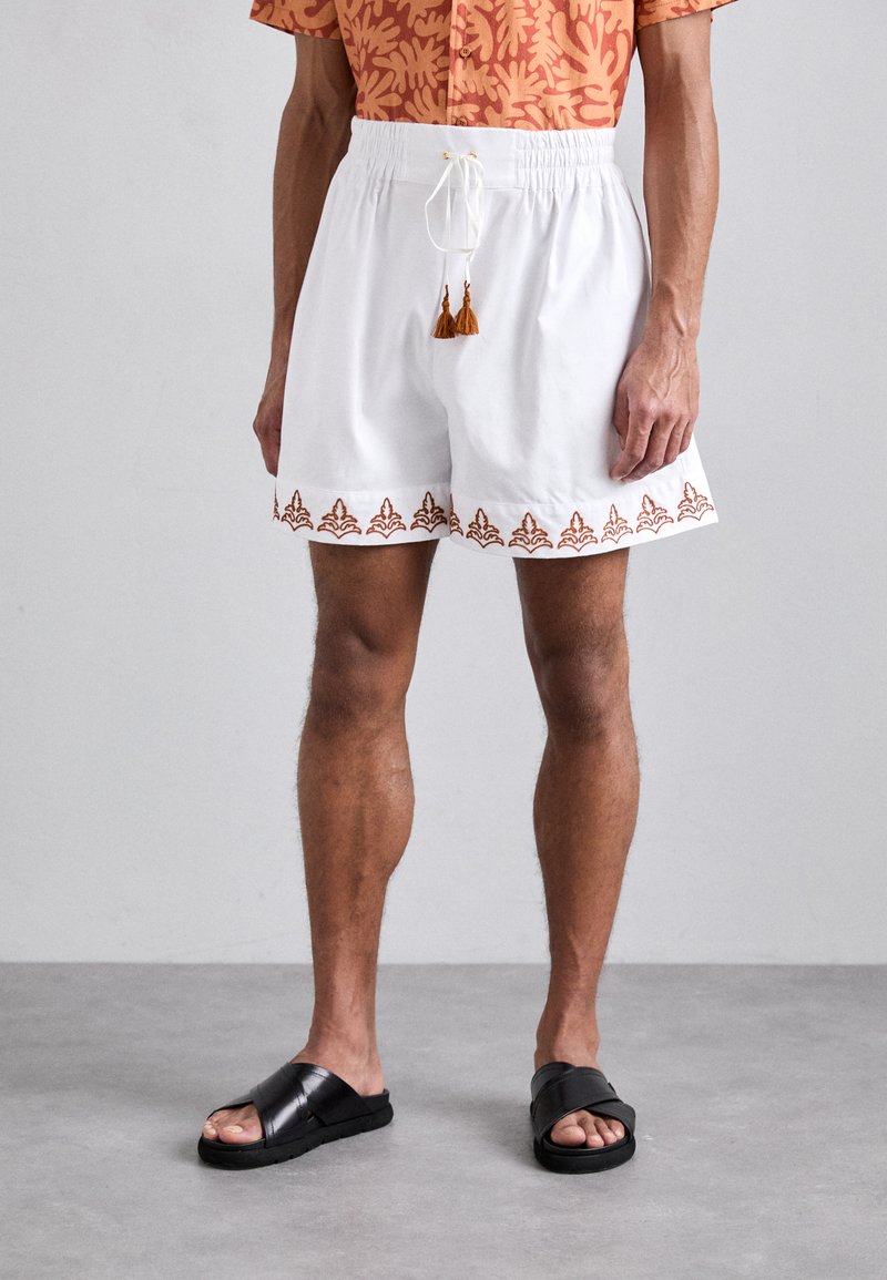 White shorts with decorative brown embroidery at the hem, featuring an elastic waistband and drawstring with tassels. Pairing with black slide sandals.