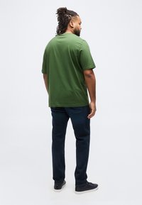 Άνδρας με dreadlocks που στέκεται γυρισμένος με την πλάτη, φοράει πράσινη μπλούζα, σκούρο μπλε τζιν και μαύρα αθλητικά παπούτσια σε λευκό φόντο.