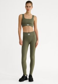 Sport-bh i olivgrönt med ribbad design och högmidjade leggings med logotags, kombinerat med svarta sportskor.