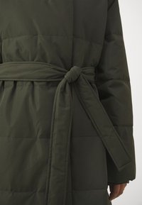 Olivově zelený prošívaný kabát s látkovým opaskem uvázaným v pase, s polstrovanou strukturou a hladkým povrchem.