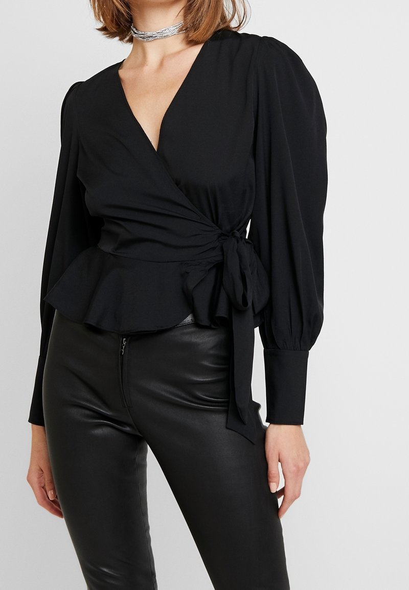 Femme portant un chemisier noir portefeuille à manches longues bouffantes et taille peplum, associé à un pantalon en cuir noir et un collier ras-du-cou argenté à plusieurs couches.