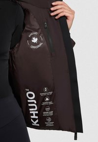Bruine geïsoleerde jas met een ritszak, met een "KHUJO ORIGINAL" logo en wasinstructies gedrukt op de binnenvoering.