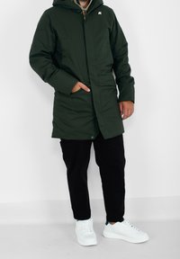 Zielona pikowana parka z zamkiem błyskawicznym i zapięciem na zatrzaski, kapturem, bocznymi kieszeniami oraz tłoczonym logo. Noszona z czarnymi spodniami i białymi butami.