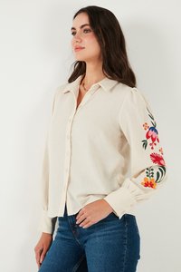 Camisa beige claro de lino con botones en el frente, que presenta un colorido bordado floral en las mangas y puños fruncidos.