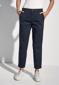 Dunkelblaue Baumwollhosen mit schmalem Schnitt, vorderen Taschen und Knopfverschluss. Stilvoll kombiniert mit weißen Sneakern, vor einem hellen Hintergrund präsentiert.