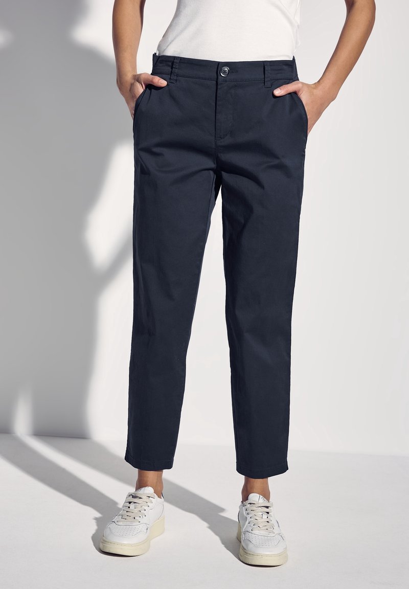 Dunkelblaue Baumwollhosen mit schmalem Schnitt, vorderen Taschen und Knopfverschluss. Stilvoll kombiniert mit weißen Sneakern, vor einem hellen Hintergrund präsentiert.