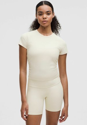 Helles beige Activewear-Set bestehend aus einem kurzärmligen Oberteil und Shorts bis zur Mitte des Oberschenkels, gefertigt aus geripptem, elastischem Stoff mit figurnahem Schnitt.