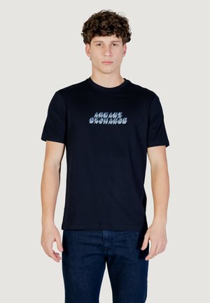 Giovane uomo con capelli ricci che indossa una t-shirt nera Armani Exchange e jeans blu scuro, in piedi contro uno sfondo chiaro e uniforme.