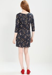 Marinblå blommig klänning med trekvartslånga ärmar och dragkedja i ryggen. Har ett fint blommönster och en figursydd siluett, kompletterad med röda skor.