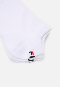 Stapel aus drei weißen Knöchelsocken mit kleinem schwarzem und rotem Logo nahe den Zehen auf hellem Hintergrund.