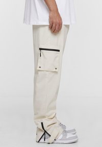 Pantaloni cargo crema con tasca laterale, dettagli in cerniera nera e caviglie con zip. Abbinati a sneakers grigie e bianche.