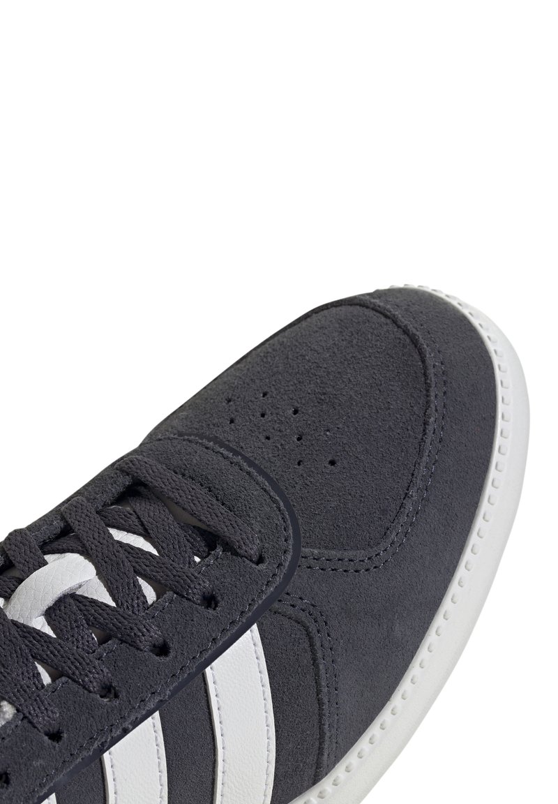 Zapatilla de ante gris con diseño de tres rayas blancas, puntera redonda, suela de goma blanca y cordones negros. Presenta detalles perforados en la parte frontal.