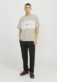 Light beige short-sleeve shirt with a white horizontal panel and printed text. Black loose-fitting trousers and brown lace-up shoes.