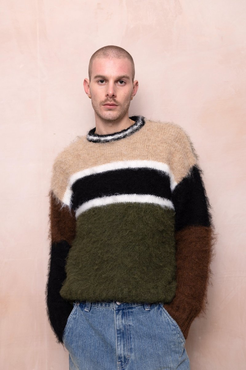 Mladenič s kratkimi lasmi, oblečen v večbarvni puhasto pulover in modre kavbojke, stoji ob svetlo rožnati steni z rokama v žepih.