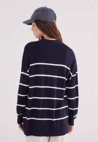 Námořnický pruhovaný svetr s bílými horizontálními pruhy, dlouhými rukávy, volným střihem, pletenou teksturou, kombinovaný s tmavou džínovou čepicí.