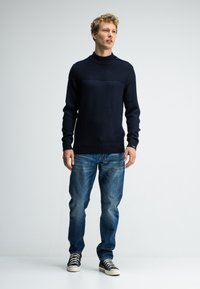 Man staat in een marineblauwe, geribbelde trui, blauwe jeans en zwarte high-top sneakers tegen een eenvoudige witte achtergrond.