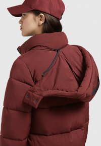 Bordo puffer jakna s visokim ovratnikom i zatvaranjem na zip. Ima kopčanje na dugme na kapuljači i mekanu, kviltanu teksturu.