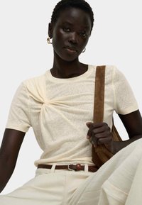T-shirt color crema con dettaglio a nodo torcigliato sul davanti, realizzato in un tessuto morbido. Il modello indossa una cintura marrone e porta una borsa in suede marrone.