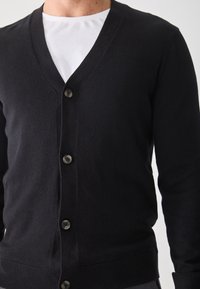 Mann, der einen schwarzen Knopf-Kardigan über einem weißen Rundhals-T-Shirt trägt, von den Schultern bis zur Taille gezeigt.