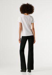 Άτομο με σγουρά μαλλιά που φοράει ένα απλό λευκό μπλουζάκι κοντό μανίκι, μαύρα τζιν και μαύρες γόβες, στέκεται στραμμένο προς τα πίσω σε λευκό φόντο.