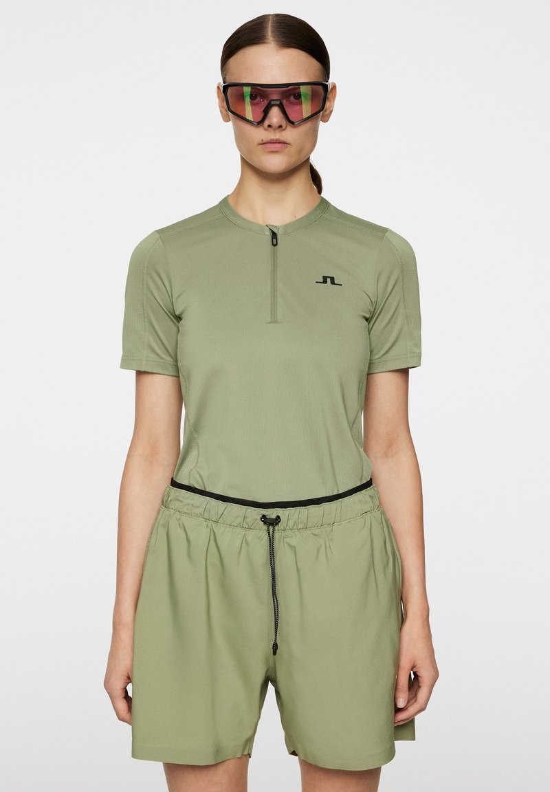 Oliivivihreä urheilupaita, jossa on puolivetoketju, lyhyet hihat ja musta logo; yhdistettynä vastaaviin rentoihin shortseihin, joissa on joustava vyötärö ja kiristysnauha.