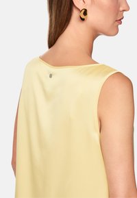 Satin yellow sleeveless top with a rounded neckline, featuring a metallic detail at the back and paired with circular black earrings.