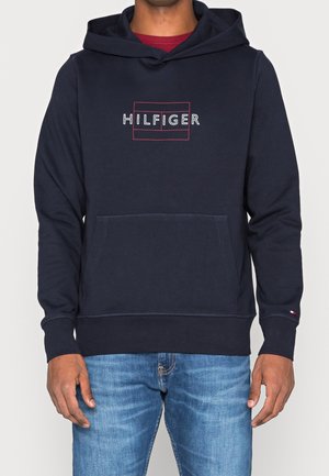 Mężczyzna ubrany w granatową bluzę z kapturem z logo „HILFIGER” z czerwonym obrysem, w jasnoniebieskich dżinsach, stojący na jednolitym tle.