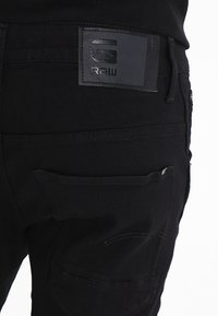 Black denim pants featuring a leather brand patch, stitched pockets, and a smooth texture. The design includes a subtle curved stitching detail.