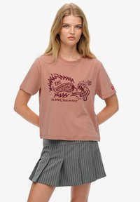 Camiseta de algodón color rosa con mangas cortas y cuello redondo, con un estampado gráfico de llamas y texto. Combinada con pantalones cortos gris con rayas finas.