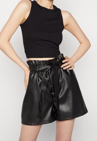 Černý žebrovaný tílko s crop střihem, kombinovaný s vysokým pasem černých kraťasů z umělé kůže s páskem na mašli a bočními kapsami.